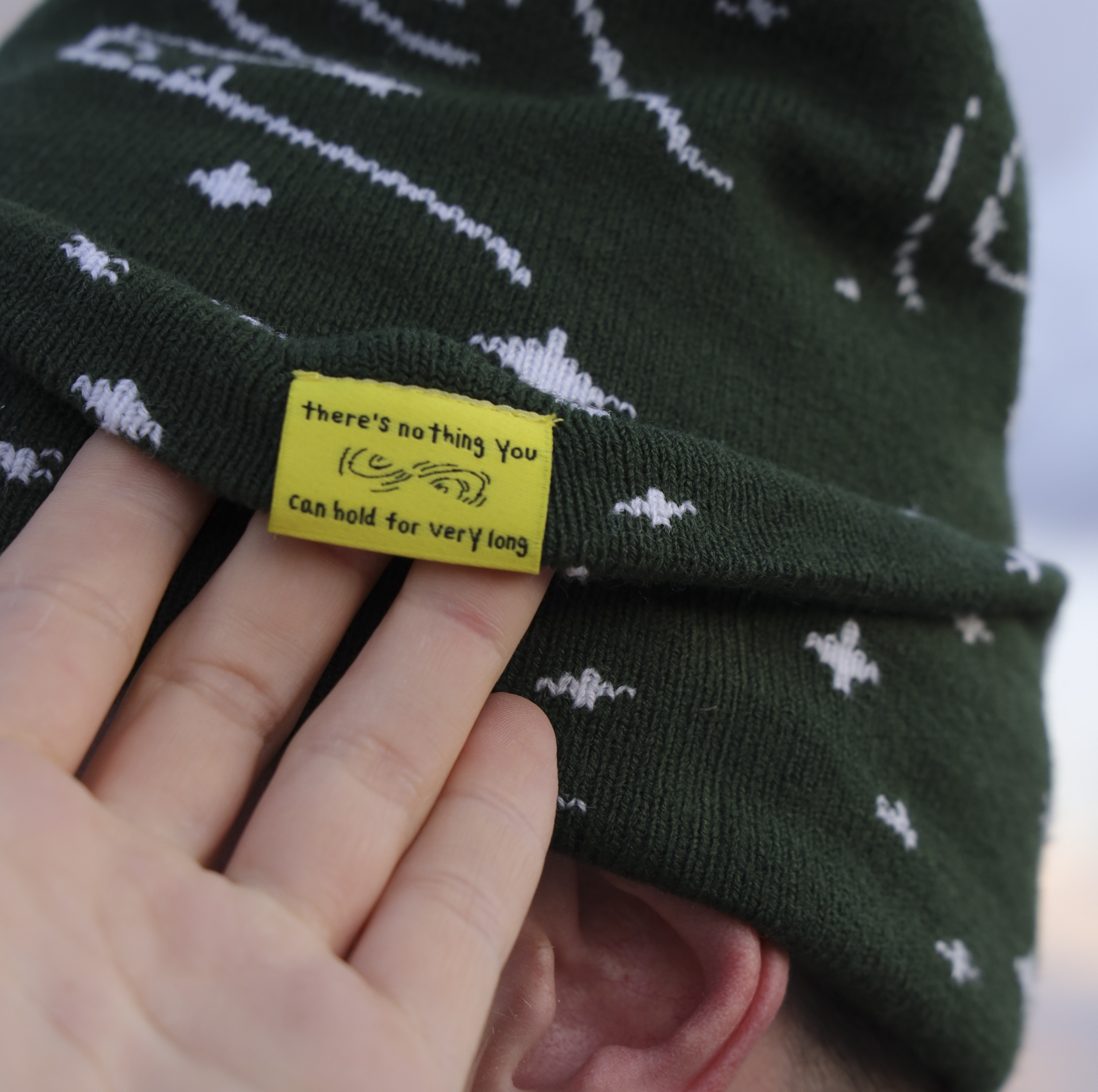 Green beanie with white patterns and a yellow tag held by a hand